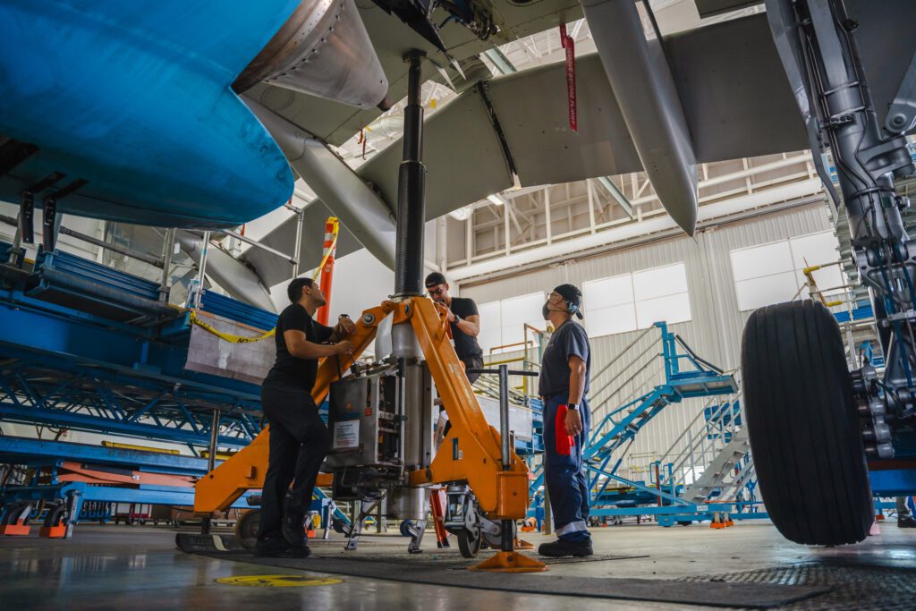 Three engineers work beneath an airplane, inspecting or repairing its underside. They wear safety gear, focused on their tasks. In the background, a hangar reveals a professional aviation maintenance environment.