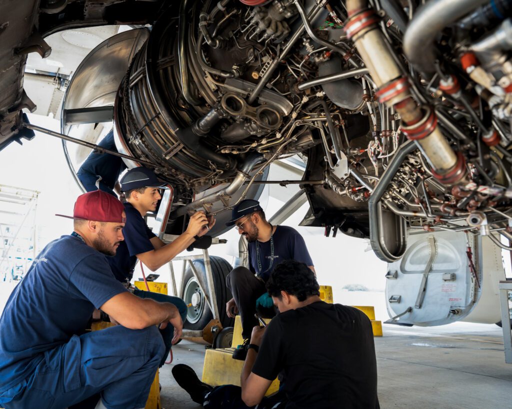 Four male workers are performing maintenance tasks on the underside of an airplane engine, working together in an aerospace facility.